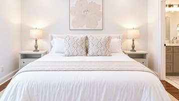 Een slaapkamer met een groot bed, twee nachtkastjes, een wandlampje en een bloemig kunstwerk boven het bed. Een badkamer is zichtbaar door een geopende deur.