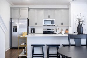 A modern kitchen with a refrigerator, microwave, coffee maker, and oven. There are two bar stools and a dining table with chairs. - The Swayze, 3BR Retreat by Velvet Ditch Villas (Oxford)