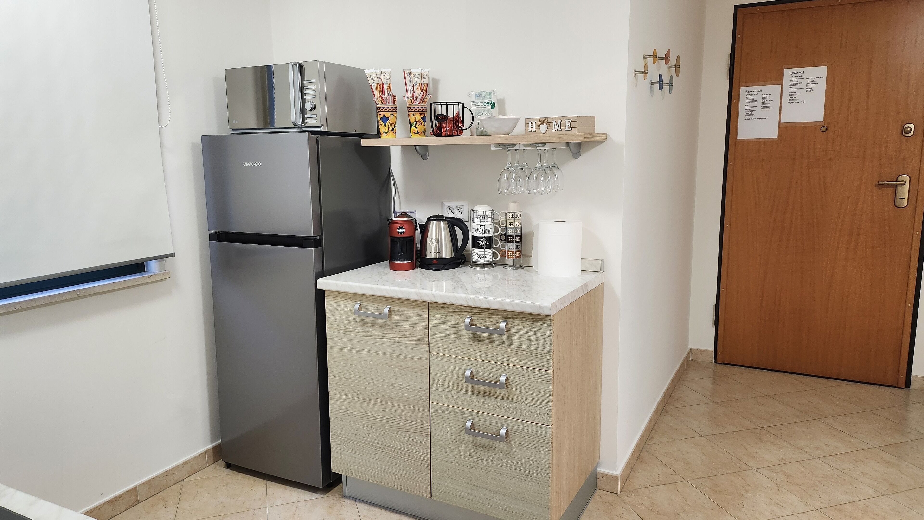 A compact kitchen with a stainless steel refrigerator, a coffee maker, and a kettle on the countertop.