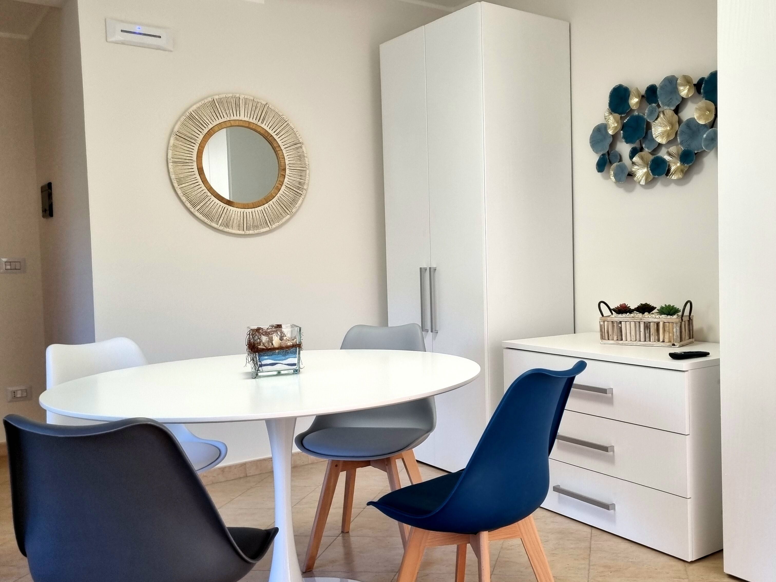 Un'area pranzo moderna con un tavolo bianco rotondo, due sedie blu, un armadietto bianco e un elemento decorativo a parete.