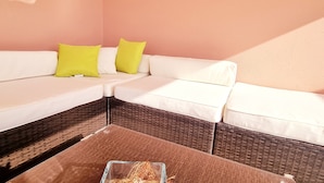 A corner sofa with white cushions and two bright yellow pillows, a wicker coffee table with a glass container, and a pink wall.