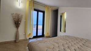 A bedroom with a large bed, a vase with dried plants, and a view of the sea through the door.