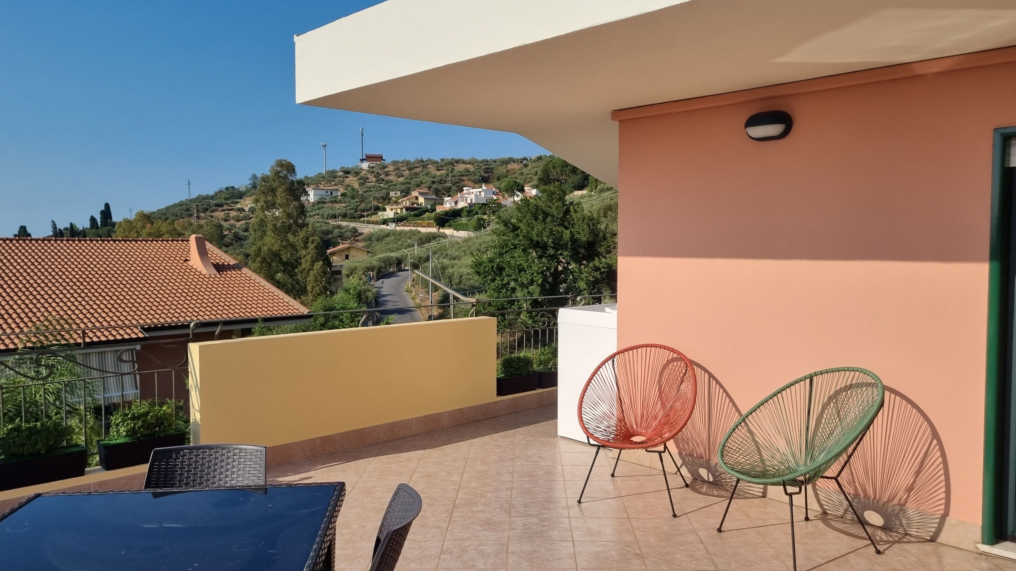 Een balkon met een betegelde vloer, twee rieten stoelen, een tafel en uitzicht op een heuvel met huizen.