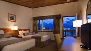 Quarto de hotel com duas camas, uma mesa de trabalho com televisão e varanda com vista para as montanhas.