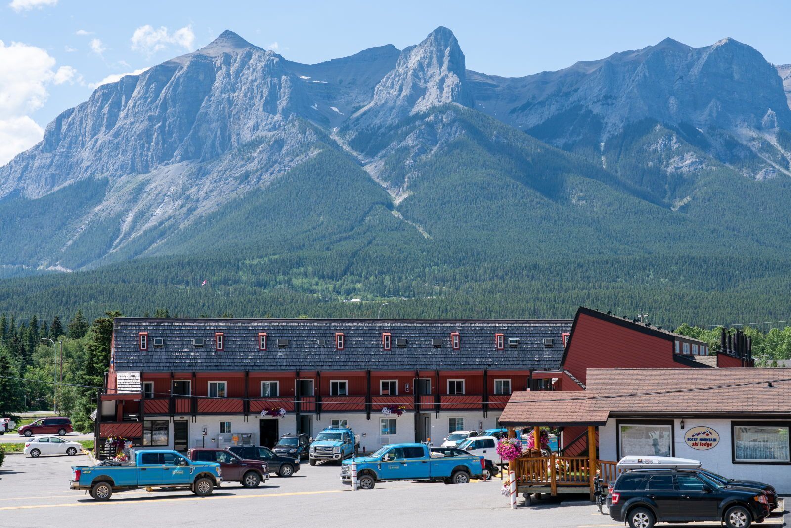 A building with a red and white facade, a parking lot with several vehicles, and a mountainous backdrop.