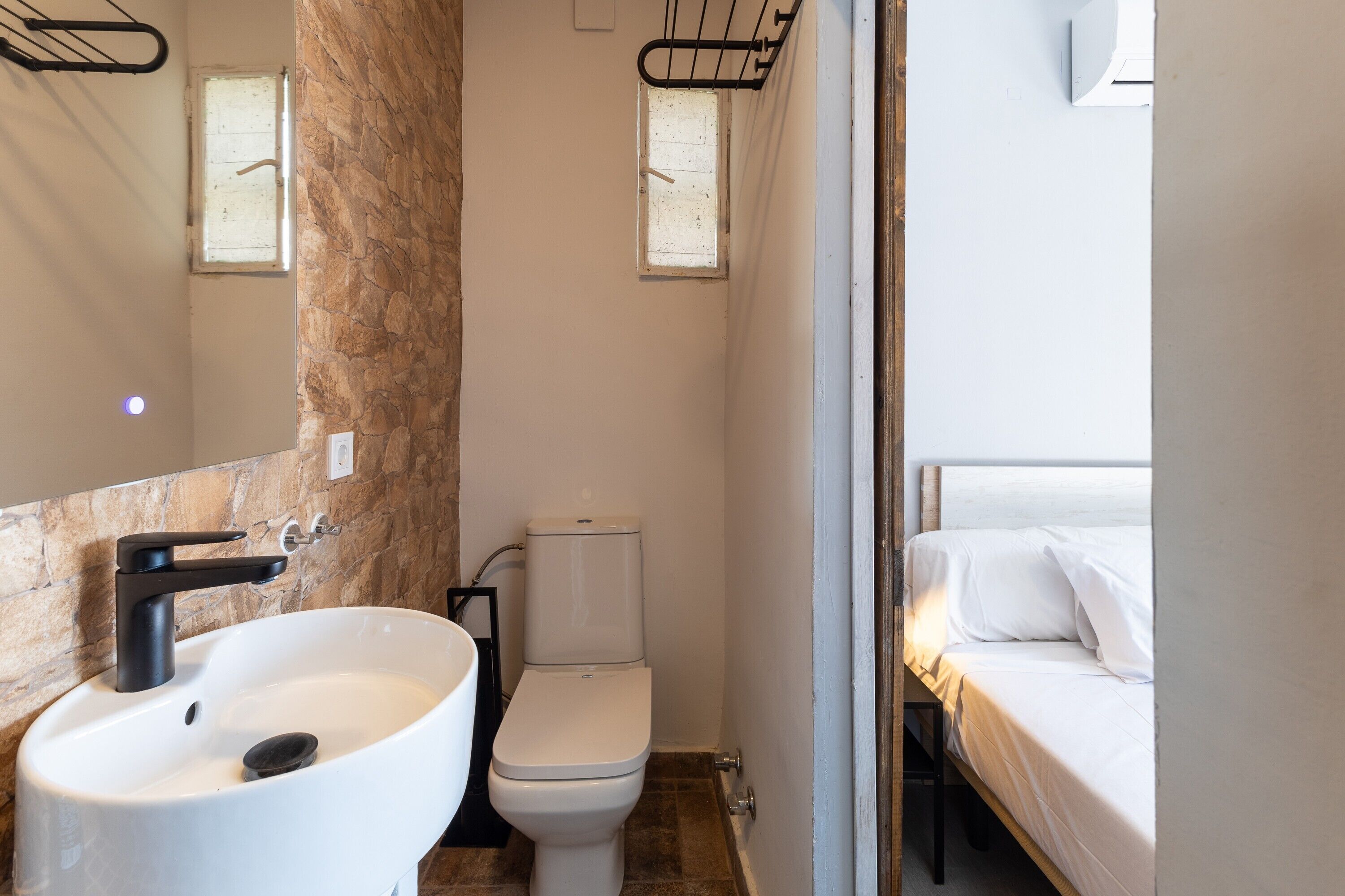 Ein Badezimmer mit Waschbecken, Toilette und einem Bett, das durch eine offene Tür sichtbar ist.