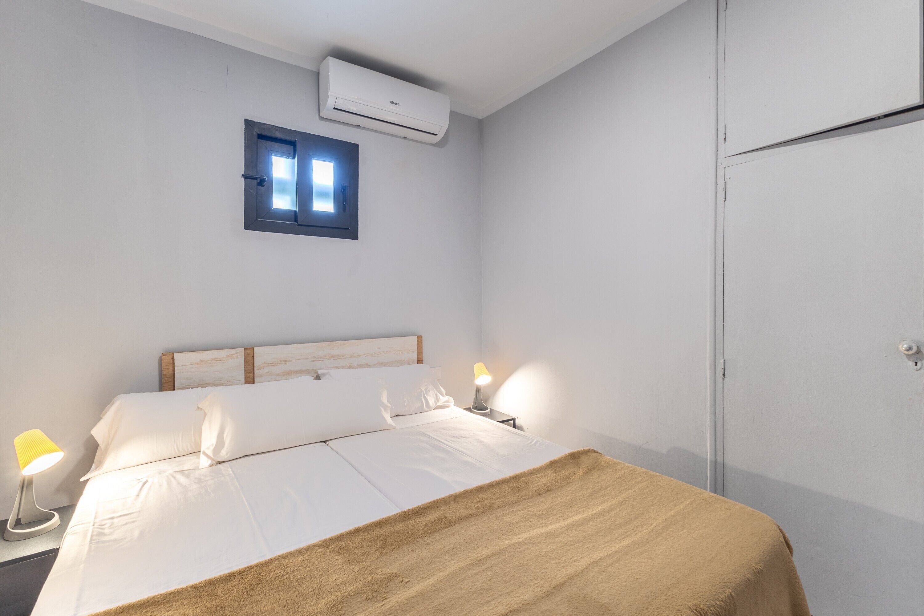 A bedroom with a bed, bedside lamps, a window, and an air conditioning unit.
