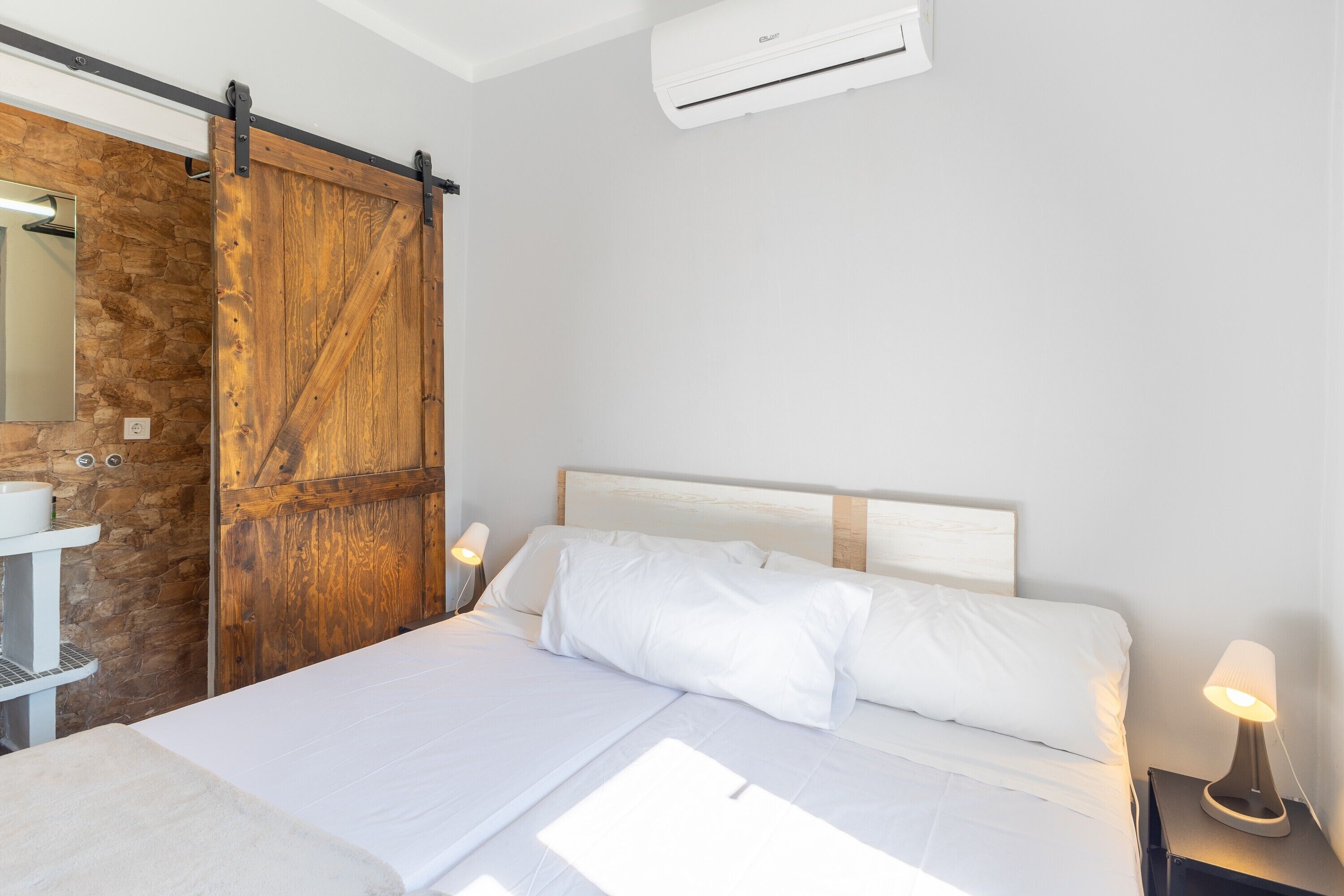 Une chambre à coucher comprenant un lit, une porte coulissante en bois, une table de chevet blanche, une lampe et un appareil de climatisation.