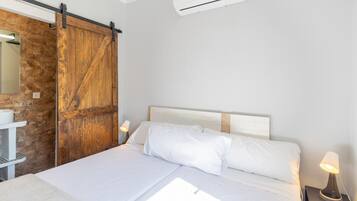 Une chambre à coucher comprenant un lit, une porte coulissante en bois, une table de chevet blanche, une lampe et un appareil de climatisation.
