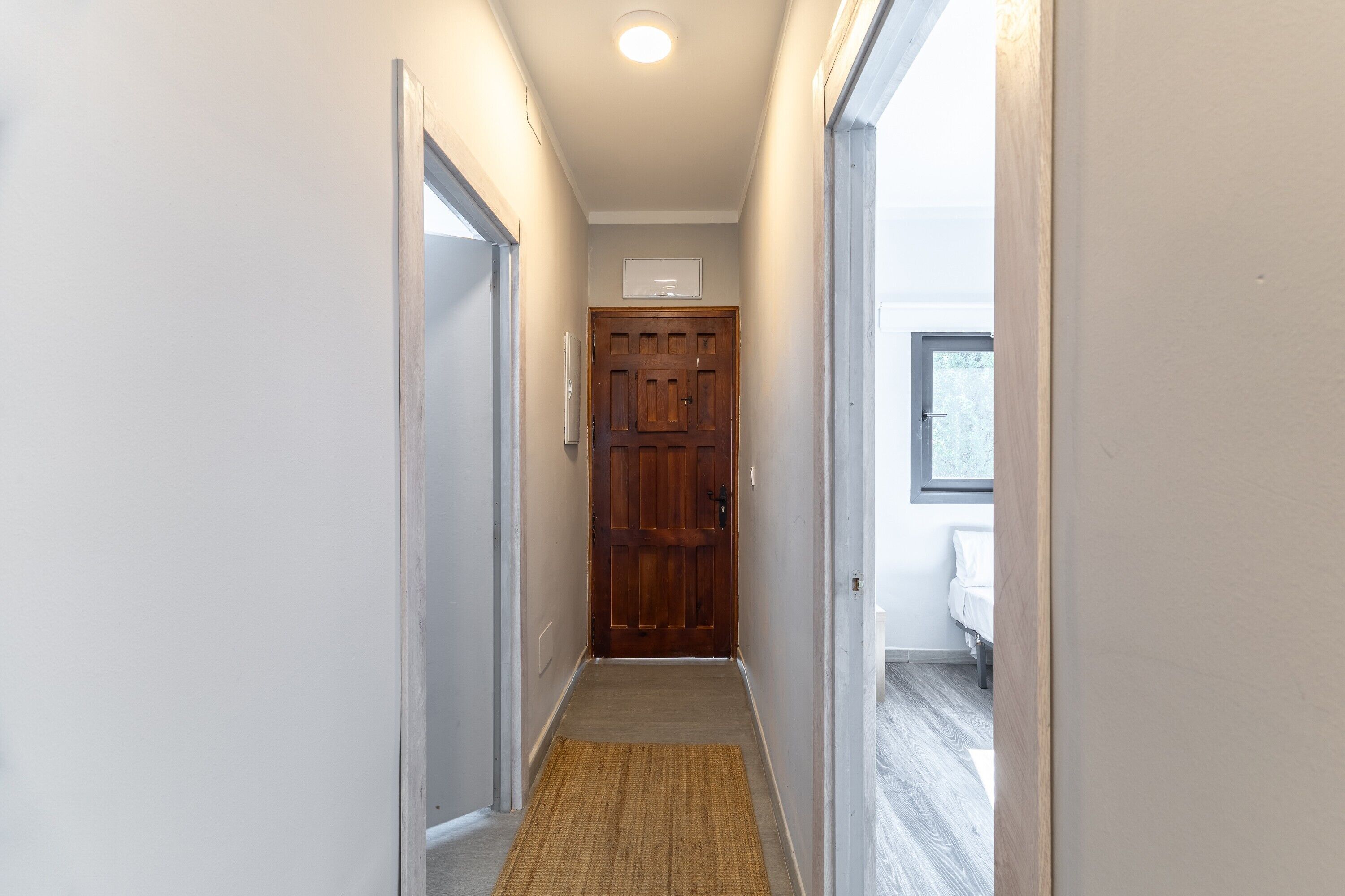 Un pasillo estrecho con una puerta de madera al final, una alfombra pequeña en el suelo y una ventana a la derecha.