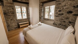 Ein Schlafzimmer mit Steinwänden, einer hölzernen Bank, einem Bett mit weißer Bettwäsche und zwei Fenstern mit Blick auf Gebäude.