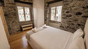 A bedroom with a stone wall, a wooden bench, a bed with white linens, and two windows with views of buildings. - Apartment in Câmara de Lobos | Privileged location (Câmara de Lobos)