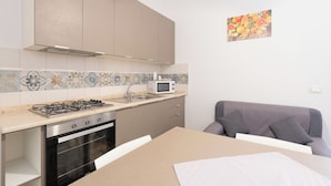 A compact kitchen with a gas stove, oven, microwave, and a sink. There is a dining table with chairs and a gray sofa. - Cozy, with garden, parking space, and 5 minutes from cornino bay beach (Sicilia)