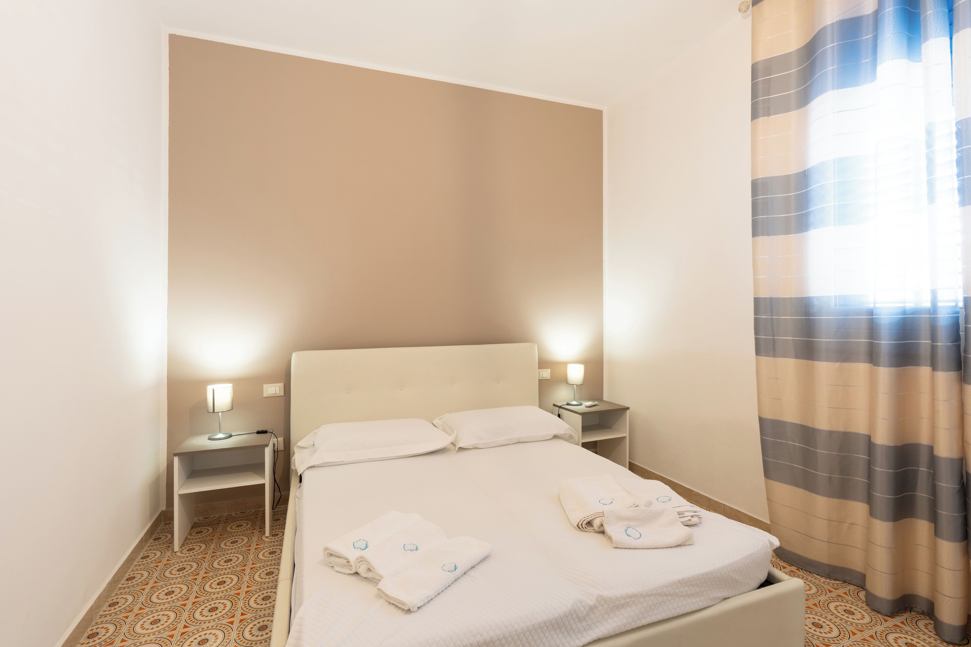 A neatly made bed with two pillows, a bedside table with a lamp, and a striped curtain.