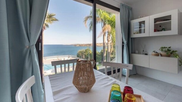 Une chambre moderne avec un balcon donnant sur la mer, comprenant une table blanche agrémentée d’un vase en osier et de verres colorés, ainsi qu’un meuble blanc avec des portes en verre.