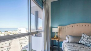 Un dormitorio con un ventanal grande, cama con ropa de cama estampada, mesita de noche con lámpara y vistas a la ciudad y al mar.