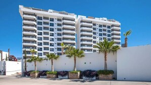Un hotel de varios pisos con balcones, fachada blanca y palmeras al frente.