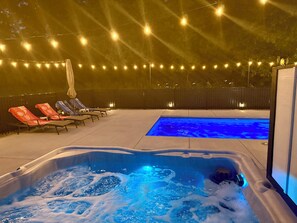 A hot tub with bubbling water, a swimming pool, lounge chairs, and string lights.