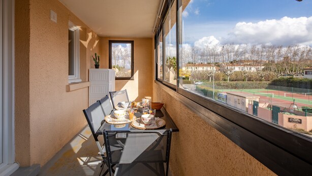 Een balkon met een ontbijttafel, een bloempot en uitzicht op een tennisbaan en gebouwen.