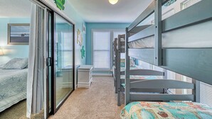 A bedroom with bunk beds, a sliding glass door, a small bedside table, and a bed with a patterned bedspread. - A Beach Escape- 1973 New River Inlet Rd (North Topsail Beach)