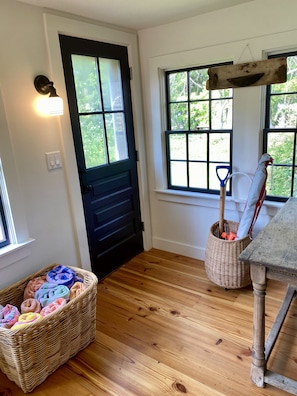 A room with a black door, a wicker basket with colorful towels, a wooden table, and a window with a view of greenery. - Little Queen - 3 Bedroom Renovated Cottage in Chatham (Chatham)