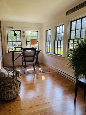 A room with a wooden floor, a chair, a table, a lamp, a basket, and a plant. - Little Queen - 3 Bedroom Renovated Cottage in Chatham (Chatham)