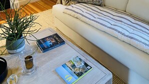 A living room with a white sofa, a small table with a plant, a book, and a glass candle holder. - Little Queen - 3 Bedroom Renovated Cottage in Chatham (Chatham)