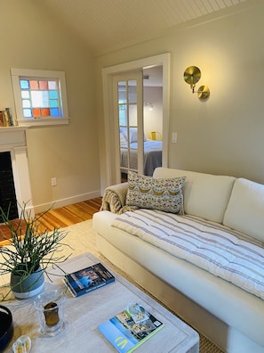 A living room with a white sofa, a small table with a plant, a book, and a glass candle holder. - Little Queen - 3 Bedroom Renovated Cottage in Chatham (Chatham)