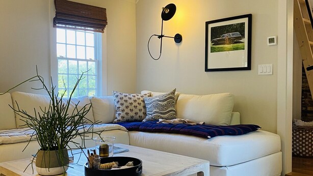 A living room with a sofa, a coffee table, a potted plant, and a wall-mounted light fixture. - Little Queen - 3 Bedroom Renovated Cottage in Chatham (Chatham)
