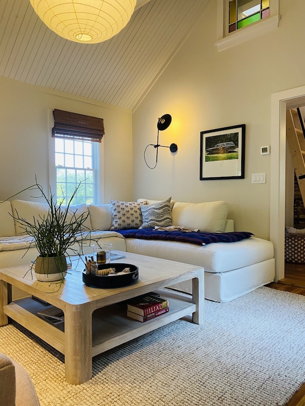 A living room with a sofa, a coffee table, a potted plant, and a wall-mounted light fixture. - Little Queen - 3 Bedroom Renovated Cottage in Chatham (Chatham)