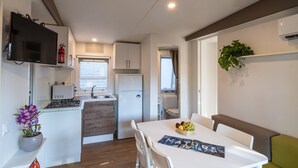 A compact kitchen with a refrigerator, stove, and sink. There is a dining table with chairs and a small plant on the wall. - Mobilhome im Ferienpark mit Grosser Poolanlage (Orbetello)