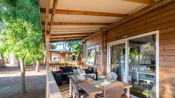 A wooden cabin with a covered patio, outdoor seating, and a sliding glass door leading to a kitchen area. - Mobilhome im Ferienpark mit Grosser Poolanlage (Orbetello)