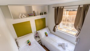 A small, clean room with two beds, a shelf with books and decorative items, and a window with curtains. - Mobilhome im Ferienpark mit Grosser Poolanlage (Orbetello)