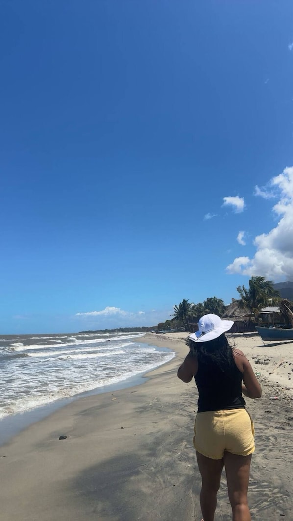 Une plage avec un ciel dégagé, une personne portant un short jaune et un chapeau blanc, et un bateau sur le sable.
