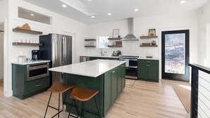A modern kitchen with a central island, green cabinets, stainless steel appliances, and a large window providing a view of the outdoors. - Prime Location! 5 Min to National Park & Wineries (Afton)