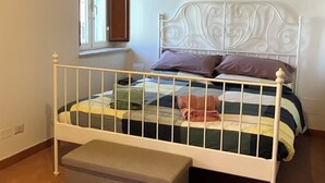 Une chambre avec un lit en métal peint en blanc, un banc au pied du lit, une fenêtre avec des volets en bois et une poutre en bois au plafond.