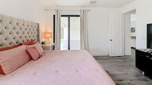 A bedroom with a tufted headboard, a large bed with pink bedding, a bedside table with a lamp, and a television on a stand. - Cozy Retreat in Port Charlotte (Port Charlotte)