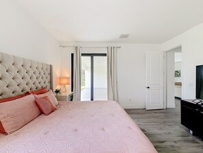 Ein Schlafzimmer mit einem gepolsterten Kopfteil, einem großen Bett mit rosa Bettwäsche, einem Nachttisch mit Lampe und einem Fernseher auf einem Schrank.