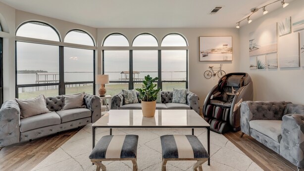 A spacious living room with a large window overlooking a body of water, a massage chair, and a sofa with a potted plant. - Gun Barrel Lakefront Retreat (Gun Barrel City)