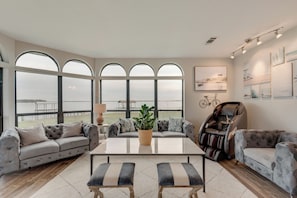 A spacious living room with a large window overlooking a body of water, a massage chair, and a sofa with a potted plant.