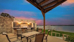 An outdoor patio with a stone wall, a wooden table, wicker chairs, and a grill area with lights. - Gun Barrel Lakefront Retreat (Gun Barrel City)
