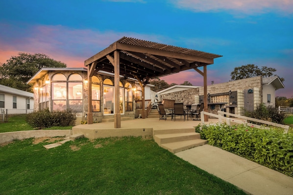 A covered patio area with a dining table and chairs, adjacent to a house with a stone facade.