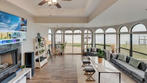 A spacious living room with a large TV, wooden flooring, a ceiling fan, and a view of the outdoors through large windows. - Gun Barrel Lakefront Retreat (Gun Barrel City)