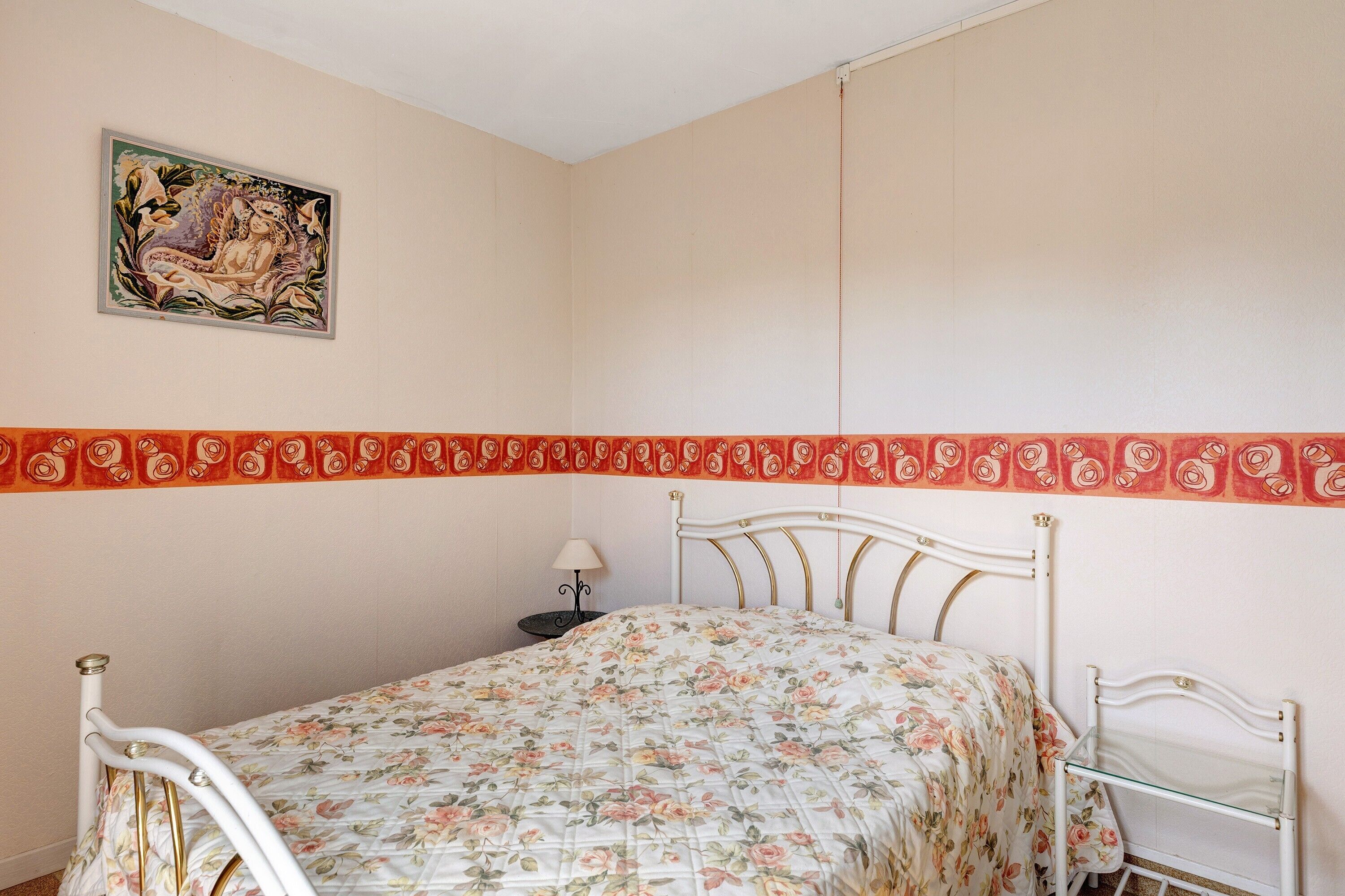 A bedroom with a bed, a nightstand, a lamp, a wall painting, and a decorative border.