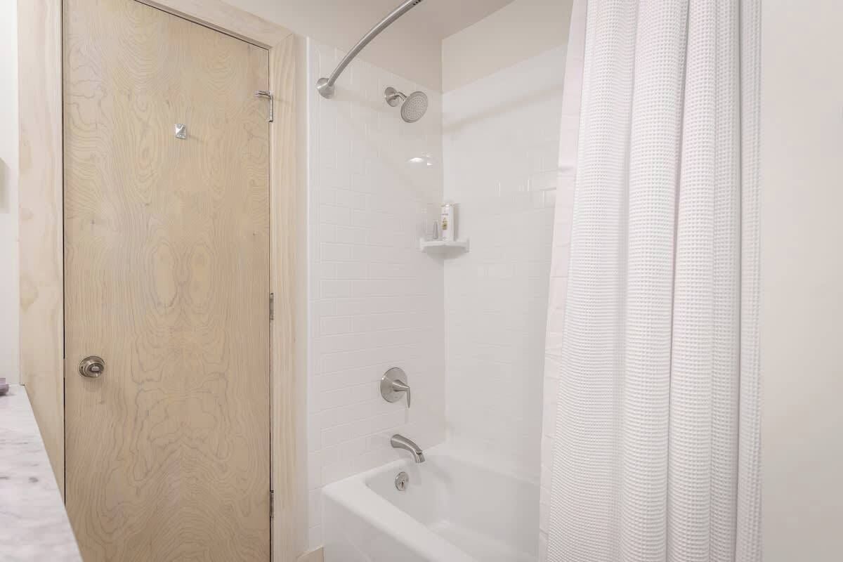 A bathroom with a wooden door, a white bathtub, a shower area, and a white tiled wall.