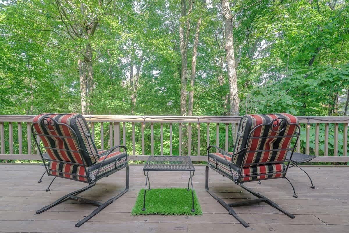 A deck with two striped chairs and a small table, surrounded by a wooden railing and lush greenery.