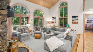 Ein geräumiges Wohnzimmer mit einem großen grauen Ecksofa, einem Couchtisch und einem Kamin. Zwei große Fenster mit Rundbögen lassen natürliches Licht herein.