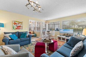 A living room with a blue sofa, a red ottoman, a TV, and a bookshelf. - Arcadian Dunes Coastal Retreat- pools, tennis, bar (Myrtle Beach)