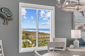 A room with a large window overlooking a beach, a chair, a table, and a lamp. - Totally Beachin' - Oceanfront, King bed, Pool (North Myrtle Beach)