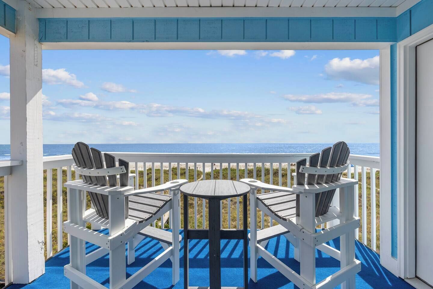 Ein überdachter Außenbereich mit zwei Adirondack-Stühlen und einem kleinen runden Tisch mit Blick auf einen Strand.